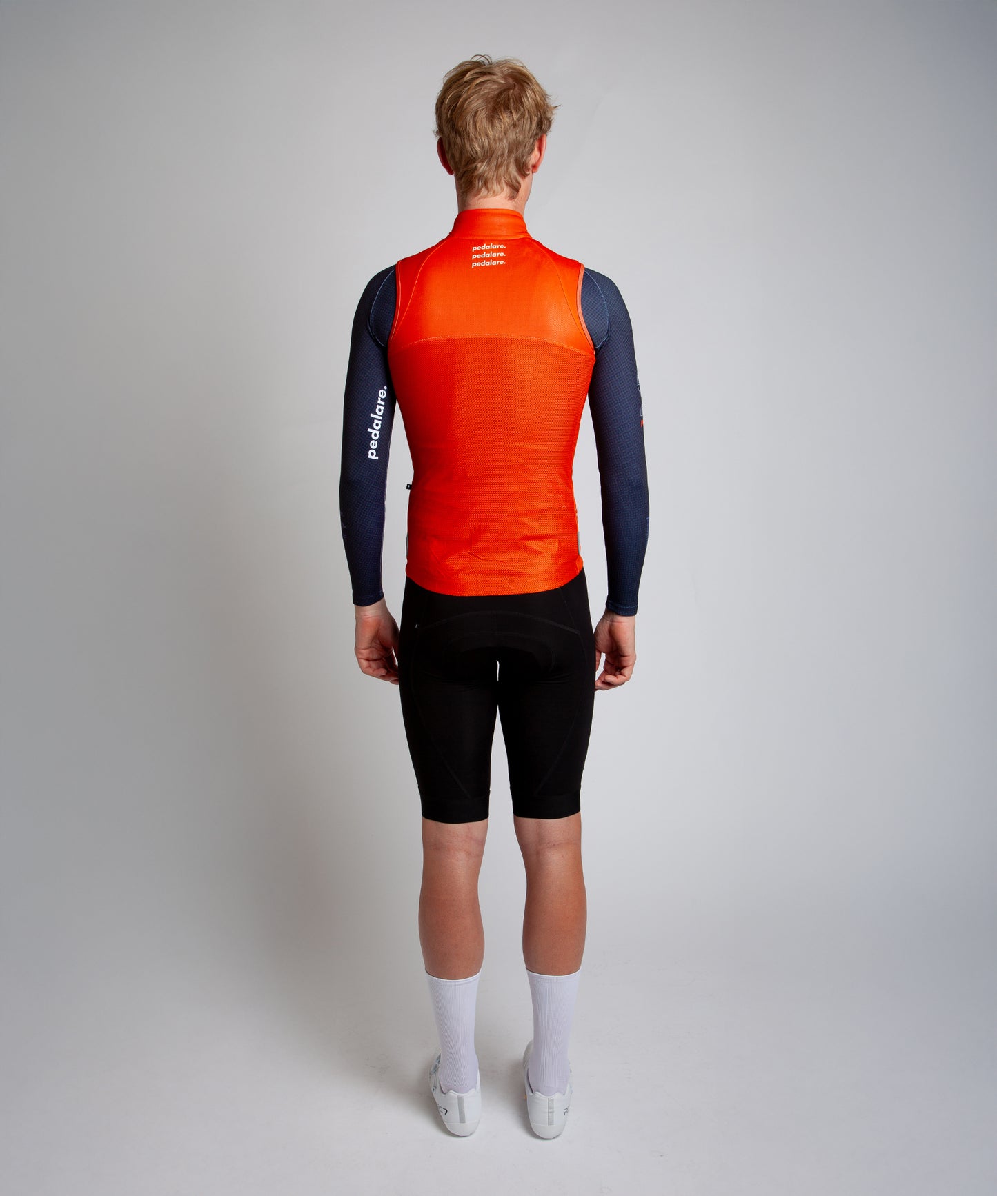 Male cyclist wearing Pedalare’s high-performance orange cycling vest, layered over a navy long sleeve jersey and black bib shorts. Side profile studio shot showcasing the vest’s aerodynamic fit and premium detailing. Lightweight, windproof men’s cycling vest ideal for cool weather rides. Designed by Perth cycling brand Pedalare for performance, comfort, and style. Aero cycling gear for dedicated road cyclists.