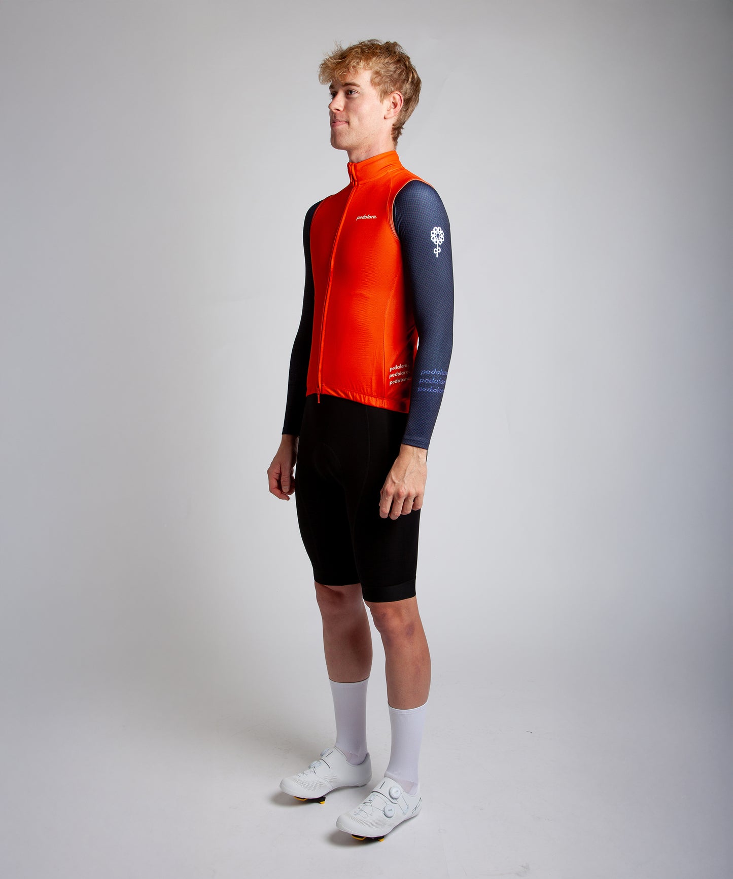 Male cyclist wearing Pedalare’s high-performance orange cycling vest, layered over a navy long sleeve jersey and black bib shorts. Side profile studio shot showcasing the vest’s aerodynamic fit and premium detailing. Lightweight, windproof men’s cycling vest ideal for cool weather rides. Designed by Perth cycling brand Pedalare for performance, comfort, and style. Aero cycling gear for dedicated road cyclists.