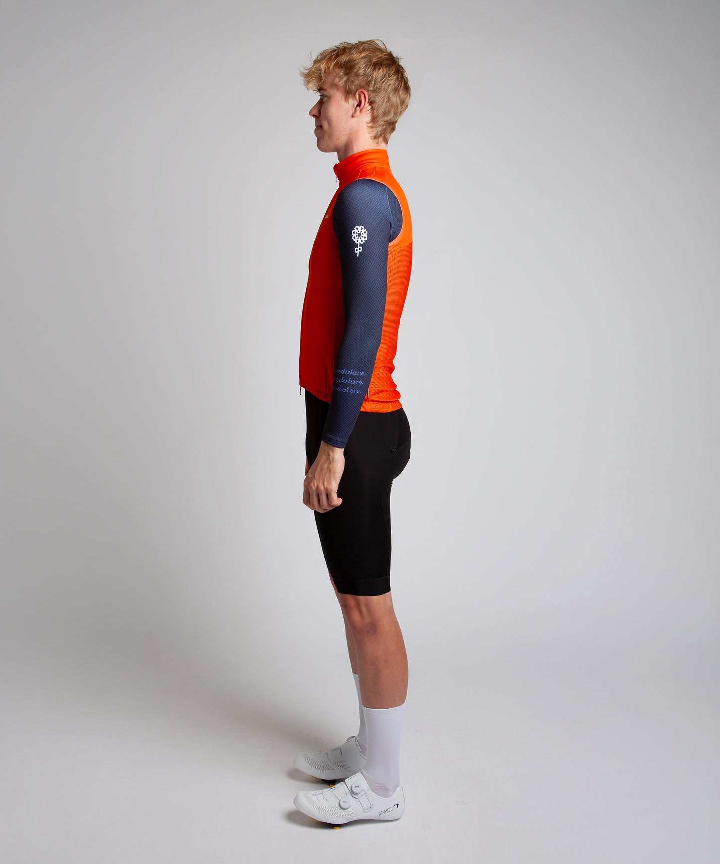 Male cyclist wearing Pedalare’s high-performance orange cycling vest, layered over a navy long sleeve jersey and black bib shorts. Side profile studio shot showcasing the vest’s aerodynamic fit and premium detailing. Lightweight, windproof men’s cycling vest ideal for cool weather rides. Designed by Perth cycling brand Pedalare for performance, comfort, and style. Aero cycling gear for dedicated road cyclists.