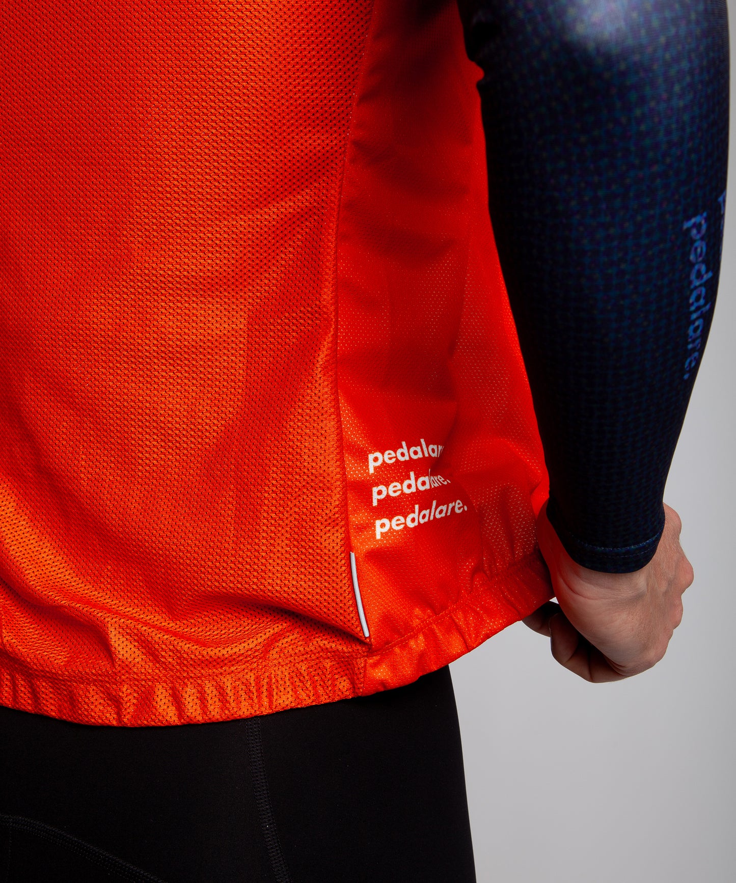 Male cyclist wearing Pedalare’s high-performance orange cycling vest, layered over a navy long sleeve jersey and black bib shorts. Side profile studio shot showcasing the vest’s aerodynamic fit and premium detailing. Lightweight, windproof men’s cycling vest ideal for cool weather rides. Designed by Perth cycling brand Pedalare for performance, comfort, and style. Aero cycling gear for dedicated road cyclists.