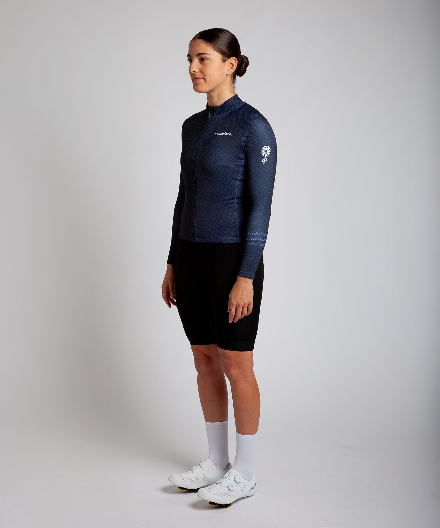 Female cyclist modelling Pedalare’s navy blue winter long sleeve cycling jersey, paired with black bib shorts. Side profile studio shot highlighting the jersey’s thermal fabric, streamlined fit, and subtle branding. Designed by Perth cycling brand Pedalare for cool-weather performance and style. Men’s winter cycling jersey ideal for road cyclists seeking aero cycling gear and premium cycling apparel.