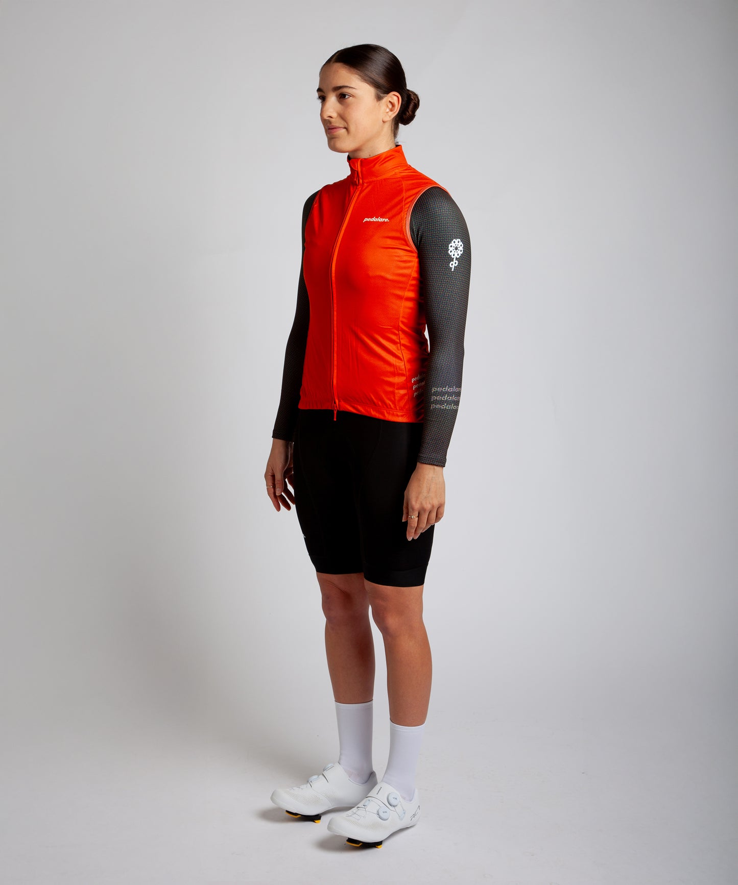 Female cyclist wearing Pedalare’s high-performance orange cycling vest, layered over a navy long sleeve jersey and black bib shorts. Side profile studio shot showcasing the vest’s aerodynamic fit and premium detailing. Lightweight, windproof men’s cycling vest ideal for cool weather rides. Designed by Perth cycling brand Pedalare for performance, comfort, and style. Aero cycling gear for dedicated road cyclists.