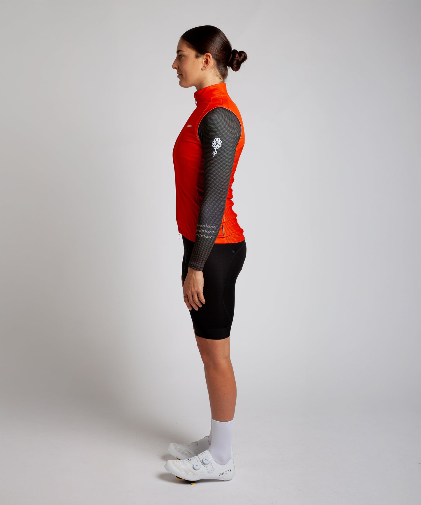 Female cyclist wearing Pedalare’s high-performance orange cycling vest, layered over a navy long sleeve jersey and black bib shorts. Side profile studio shot showcasing the vest’s aerodynamic fit and premium detailing. Lightweight, windproof men’s cycling vest ideal for cool weather rides. Designed by Perth cycling brand Pedalare for performance, comfort, and style. Aero cycling gear for dedicated road cyclists.