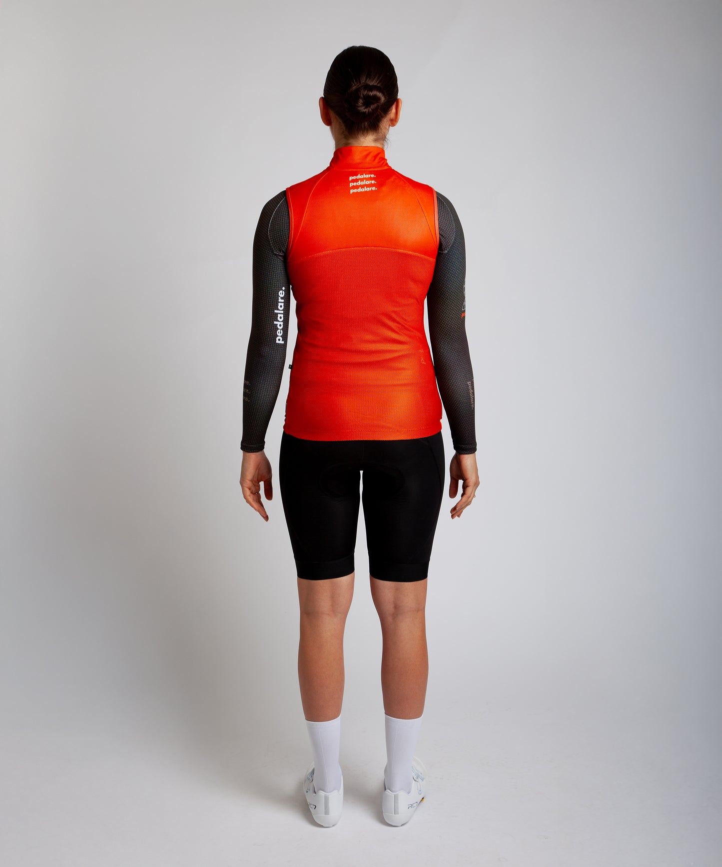 Female cyclist wearing Pedalare’s high-performance orange cycling vest, layered over a navy long sleeve jersey and black bib shorts. Side profile studio shot showcasing the vest’s aerodynamic fit and premium detailing. Lightweight, windproof men’s cycling vest ideal for cool weather rides. Designed by Perth cycling brand Pedalare for performance, comfort, and style. Aero cycling gear for dedicated road cyclists.