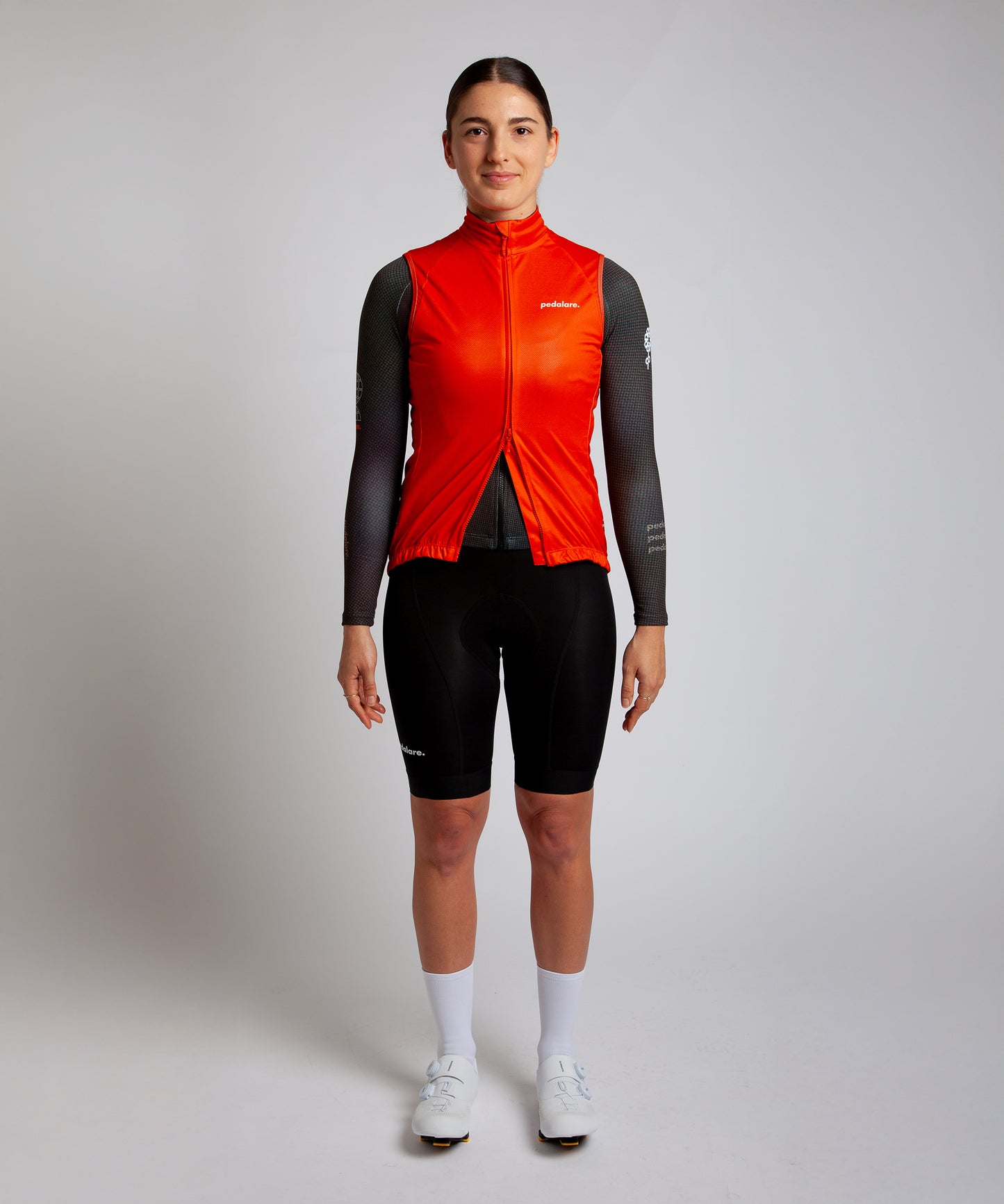 Female cyclist wearing Pedalare’s high-performance orange cycling vest, layered over a navy long sleeve jersey and black bib shorts. Side profile studio shot showcasing the vest’s aerodynamic fit and premium detailing. Lightweight, windproof men’s cycling vest ideal for cool weather rides. Designed by Perth cycling brand Pedalare for performance, comfort, and style. Aero cycling gear for dedicated road cyclists.
