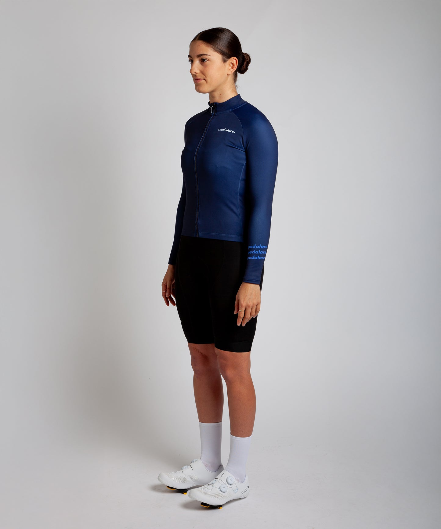 Female cyclist modelling Pedalare’s navy blue winter long sleeve cycling jersey, paired with black bib shorts. Side profile studio shot highlighting the jersey’s thermal fabric, streamlined fit, and subtle branding. Designed by Perth cycling brand Pedalare for cool-weather performance and style. Men’s winter cycling jersey ideal for road cyclists seeking aero cycling gear and premium cycling apparel.