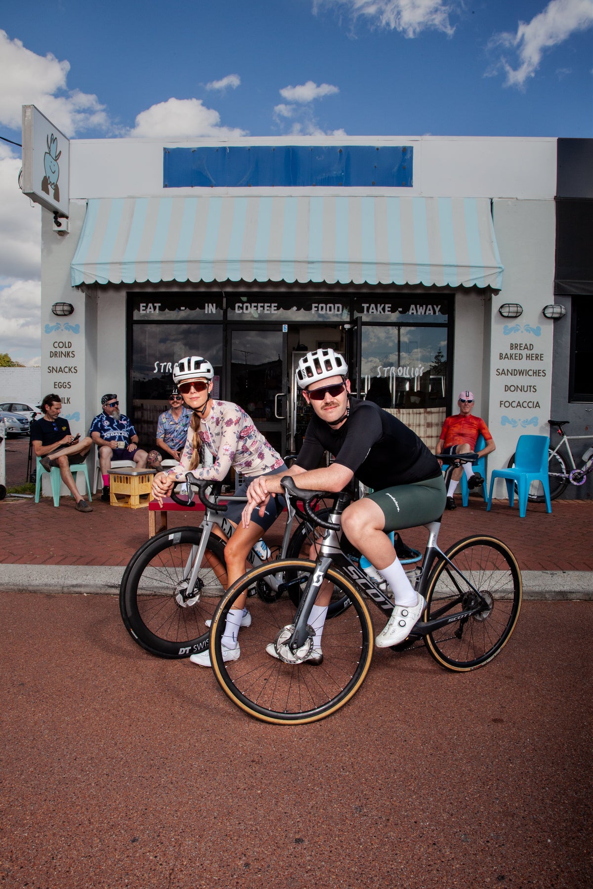 Two cyclists wearing helmets and sunglasses pose with their bicycles in front of a small cafe on a red paved street under a blue sky.