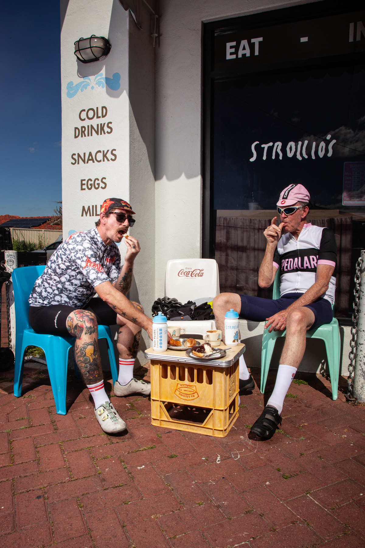 Two cyclists sitting on blue plastic chairs on a brick patio outside a cafe, with a small table between them holding drinks and snacks. A white wall on the left has printed text reading 'COLD DRINKS SNACKS EGGS MIL', and a storefront window on the right has 'STROLLO'S' written on it.