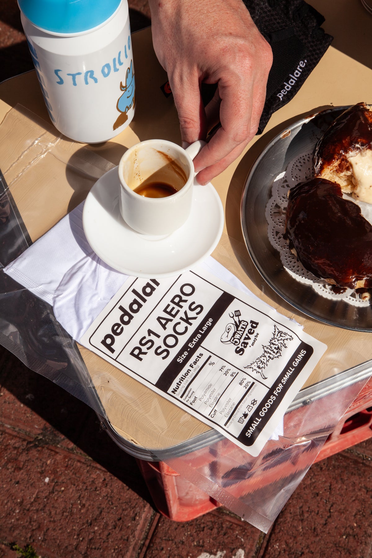 A close-up lifestyle shot of a person's hand holding a small espresso cup on a white saucer, set on a cardboard table alongside a water bottle, a chocolate-covered pastry on a metal plate, and a printed paper bag labeled 'Pedalare RS1 AERO SOCKS'.