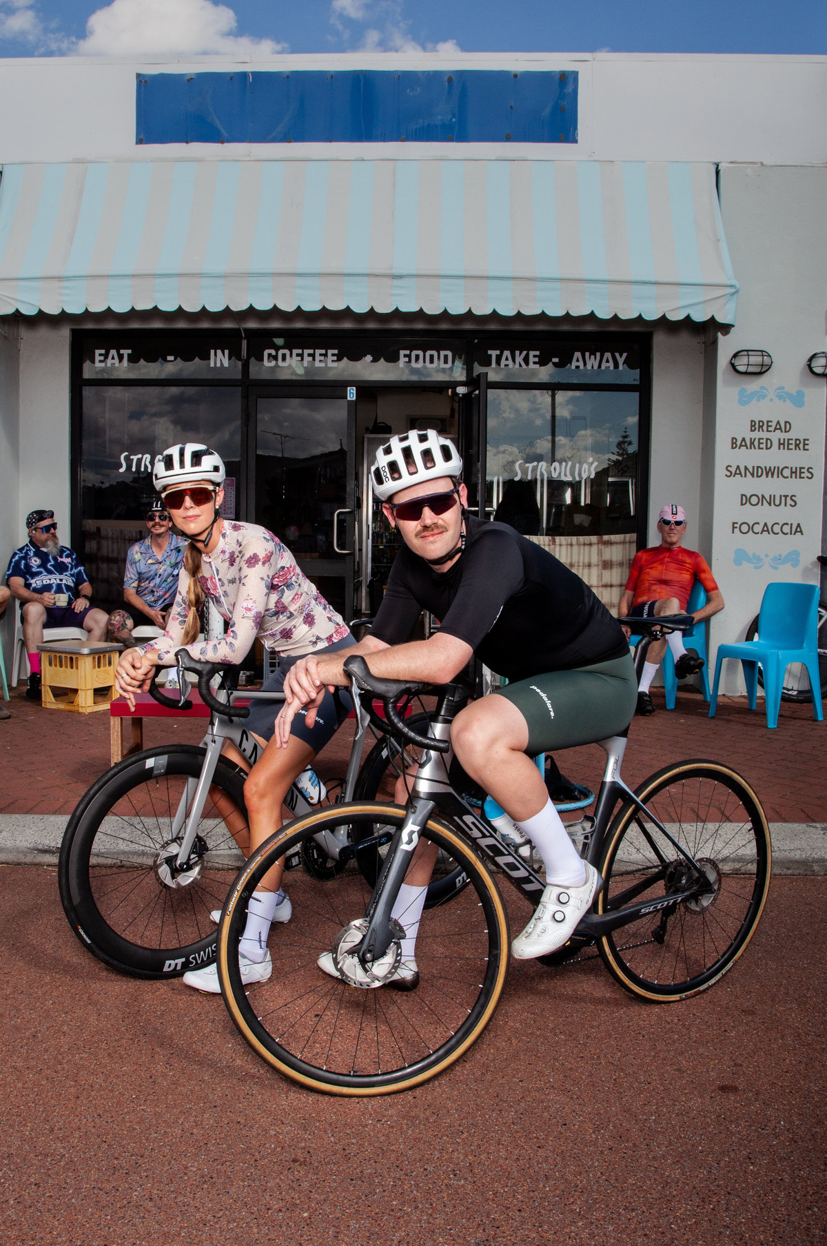 Two cyclists wearing helmets and sunglasses pose with their bicycles in front of a small cafe on a red paved street under a blue sky.
