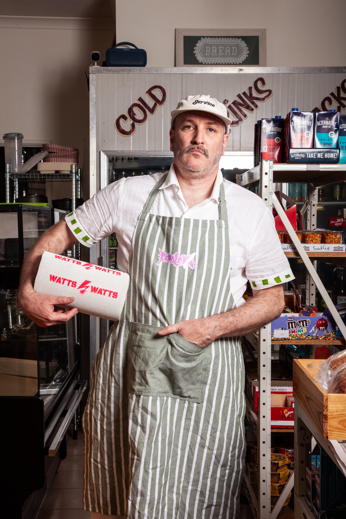 A man wearing a white shirt, a green and white striped apron, and a white cap stands in a shop holding a white box with red 'WATTS WATTS' branding. Shelves with various items and a 'COLD Drinks' sign are visible in the background.