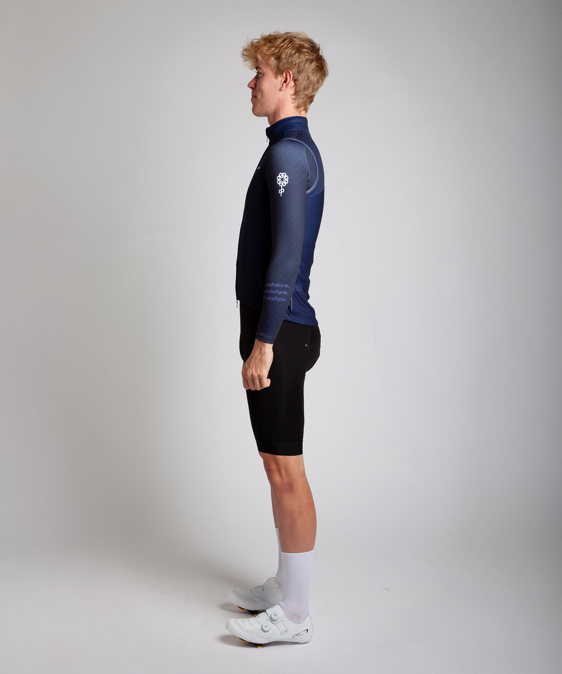 Male men’s cyclist wearing Pedalare’s high-performance navy, blue cycling vest, layered over a navy long sleeve jersey and black bib shorts. Side profile studio shot showcasing the vest’s aerodynamic fit and premium detailing. Lightweight, windproof men’s cycling vest ideal for cool weather rides. Designed by Perth cycling brand Pedalare for performance, comfort, and style. Aero cycling gear for dedicated road cyclists.