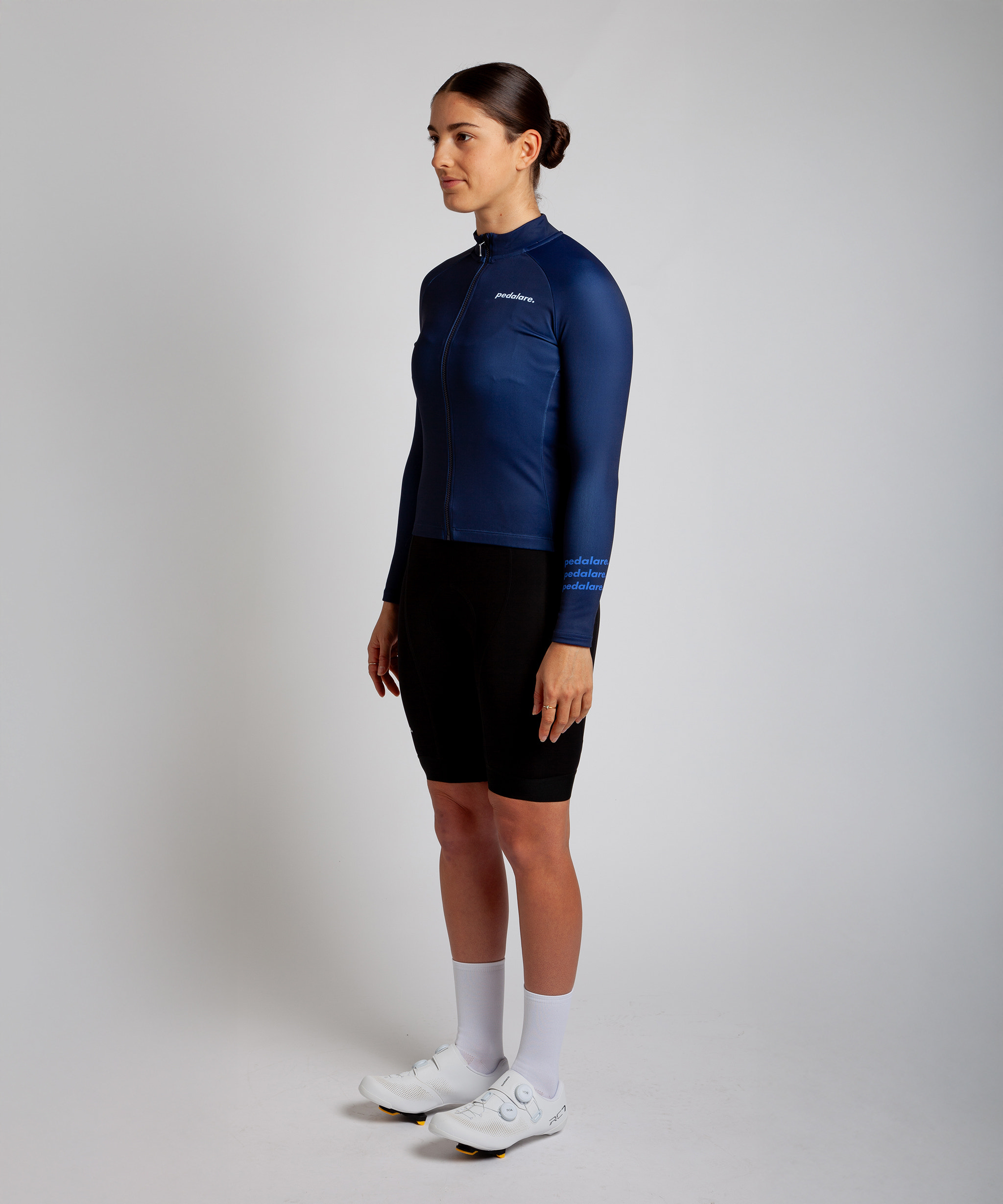 Female cyclist modelling Pedalare’s navy blue winter long sleeve cycling jersey, paired with black bib shorts. Side profile studio shot highlighting the jersey’s thermal fabric, streamlined fit, and subtle branding. Designed by Perth cycling brand Pedalare for cool-weather performance and style. Men’s winter cycling jersey ideal for road cyclists seeking aero cycling gear and premium cycling apparel.